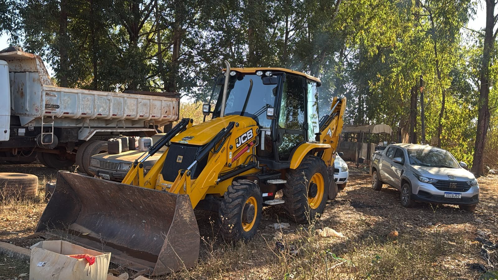 Retroescavadeira JCB parada em terreno com árvores.